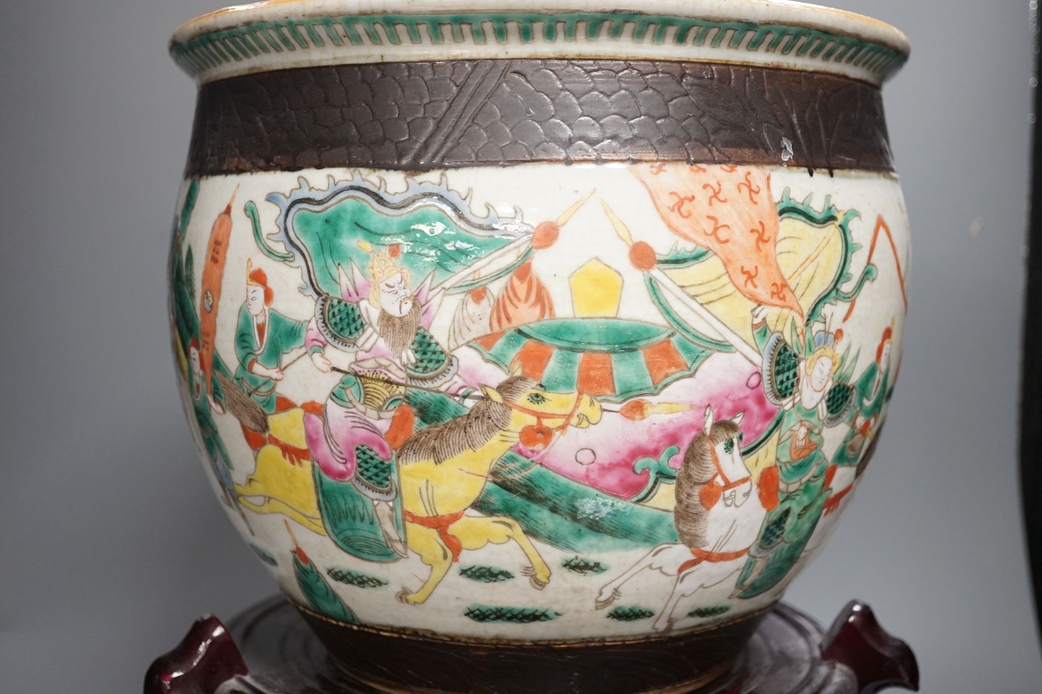 A Chinese famille rose ‘warriors’ jardiniere, late 19th century, wooden stand, 42 cms high including stand.